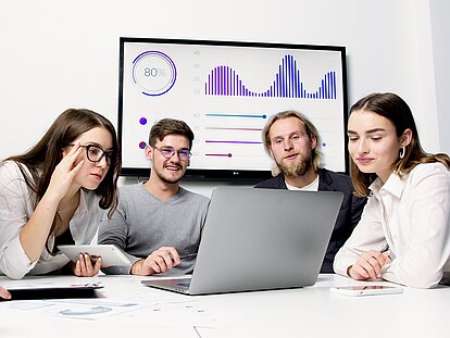 Fünf Personen blicken auf einen Laptop, im Hintergrund ist ein Fernseher zu sehen, auf dem Diagramme angezeigt werden