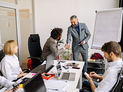 In einem Meetingraum mit fünf Personen und Laptops auf dem Tisch geben sich zwei Männer vorne am Tisch die Hand